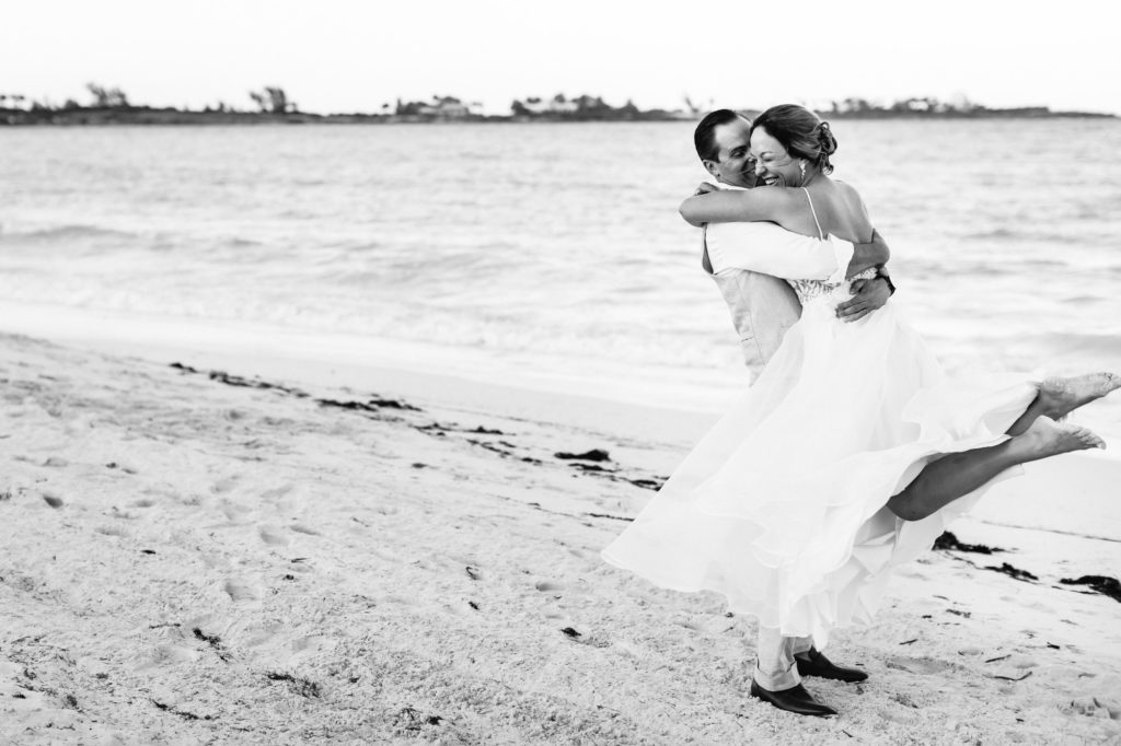 groom lifting bride up and spinning her around on the beach as newly married couple