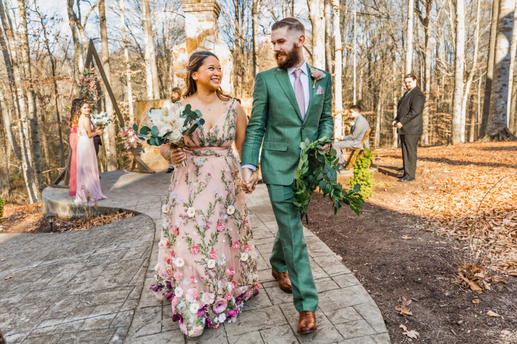 newly married couple walking together while groom wears green suit and bride wears blush pink floral embroidered wedding dress