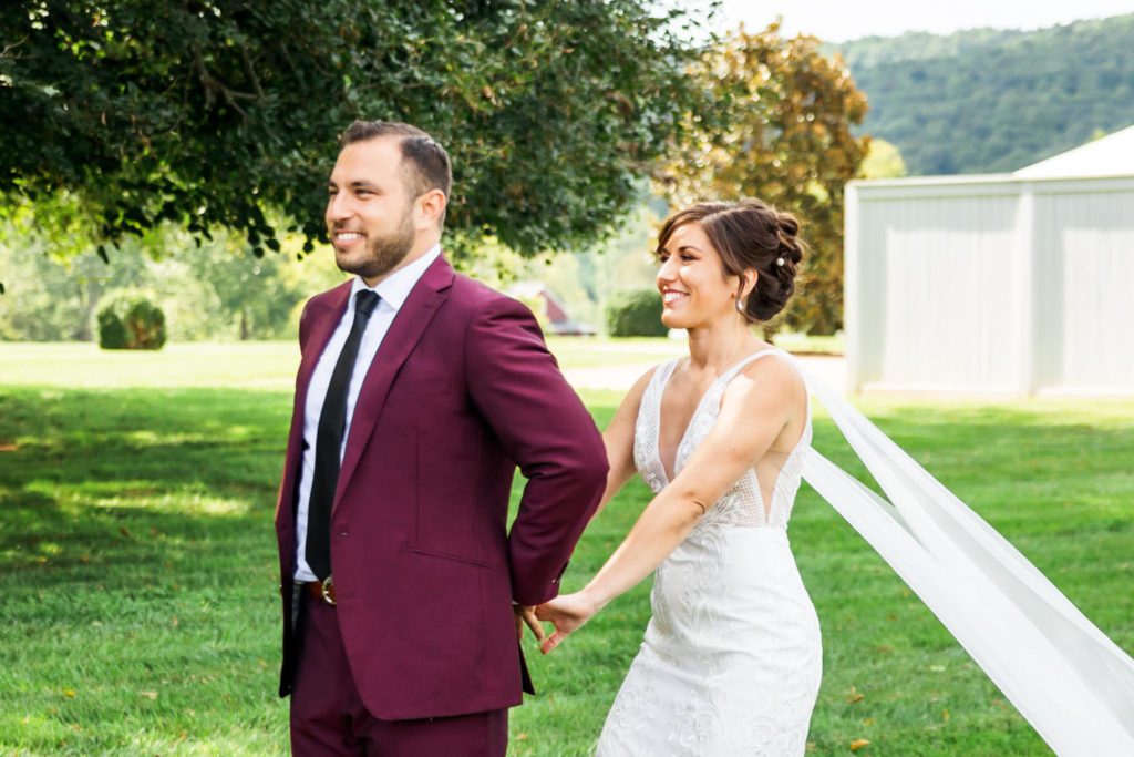 bride walking up to groom during first look wearing v-neck gown and groom wearing burgundy suit