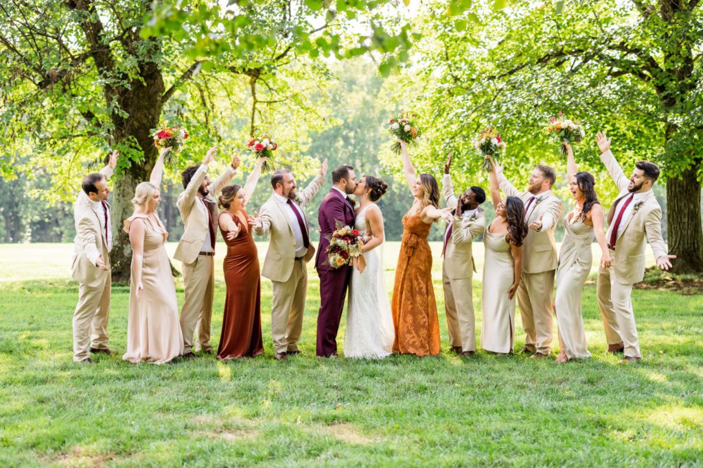 wedding party wearing neutrals and rust gowns celebrate bride and groom kissing at fall castle hill cider wedding
