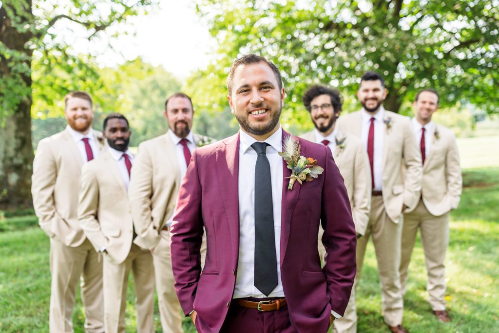 groom wearing burgundy suit smiling with groomsmen in cream suits behind him