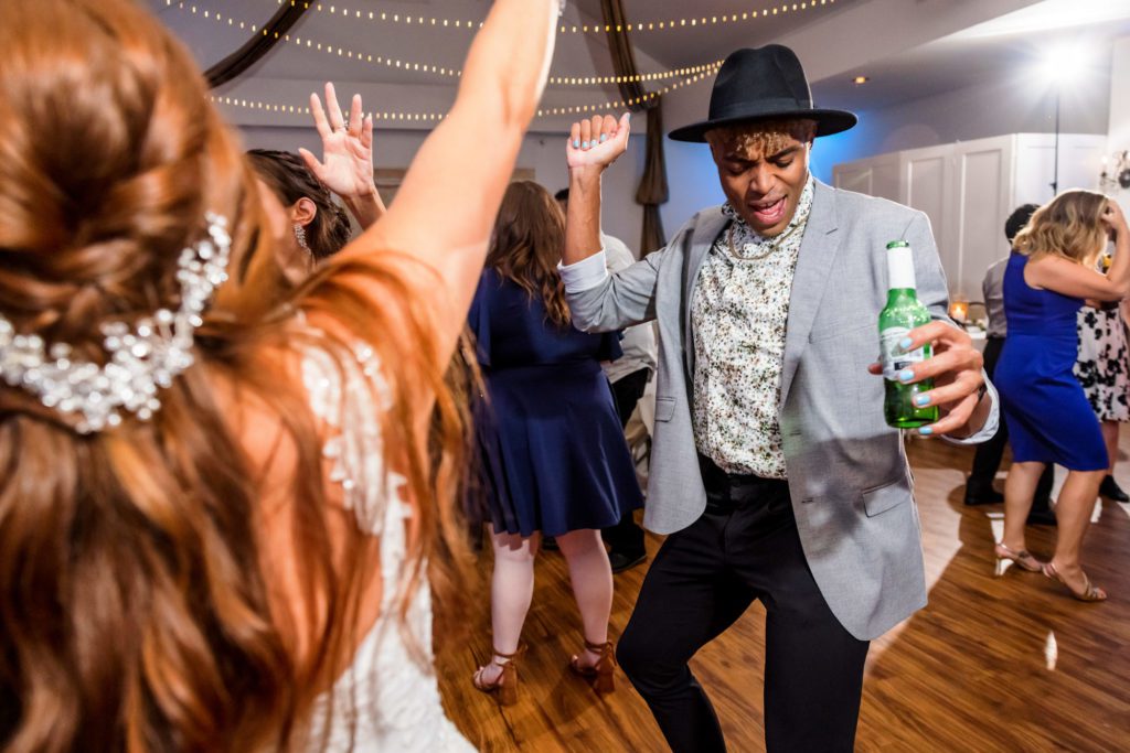 wedding guest holding beer wearing black hat dancing with bride at summer potomac point winery wedding reception