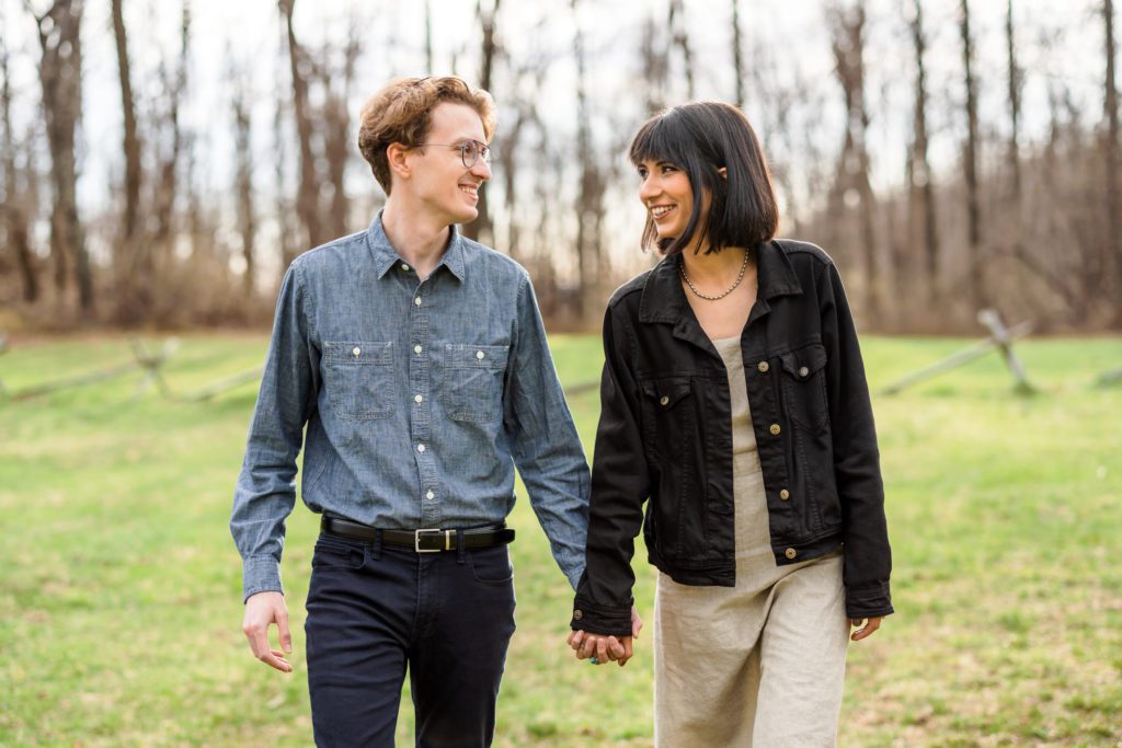 newly engaged couple walking through grass wearing casual chic engagement outfits
