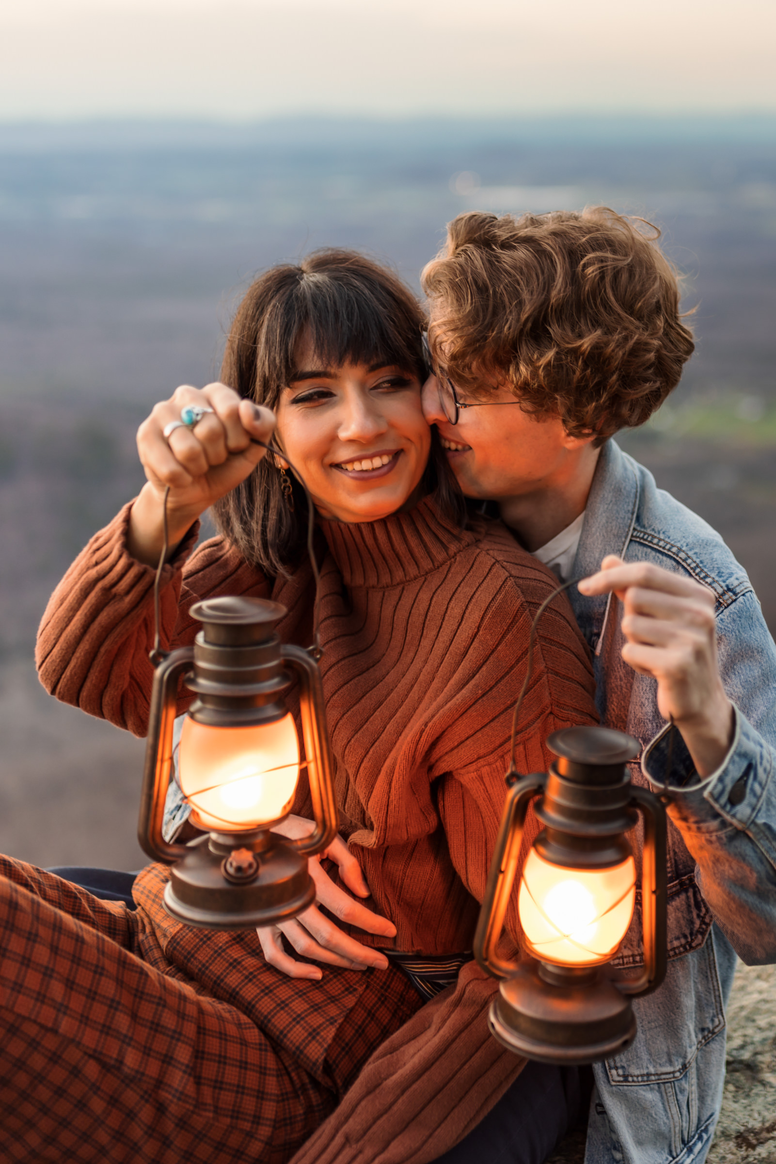 man kissing partners cheek while holding lanterns at sunset 