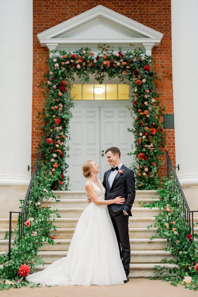 bride and groom standing together in front of stairs with flowers and greenery siding the staircase