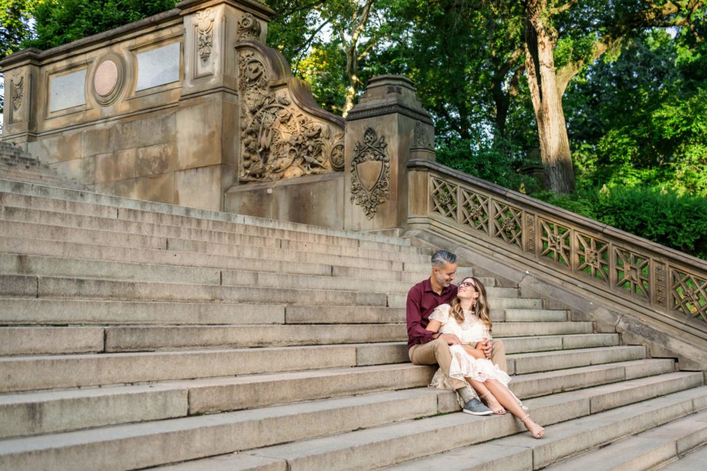 man wearing burgundy shirt and woman wearing blush dress sitting on Bethesda stairs in Central Park