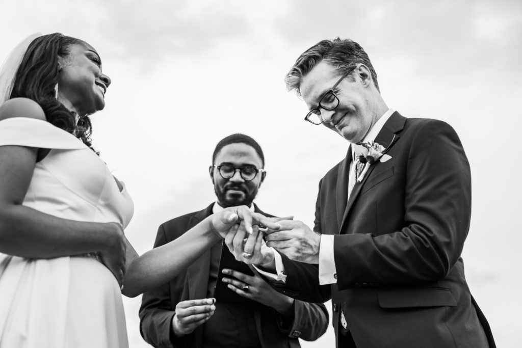 groom putting wedding band on bride's finger during outdoor ceremony