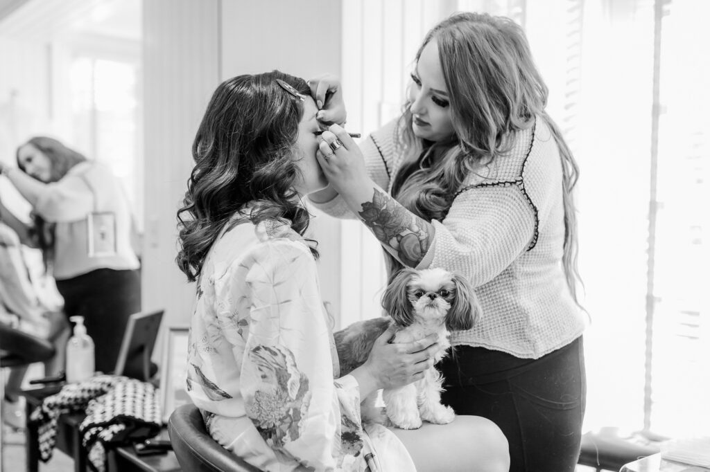 bride getting makeup done before wedding day holding dog while makeup artist puts on eyelashes
