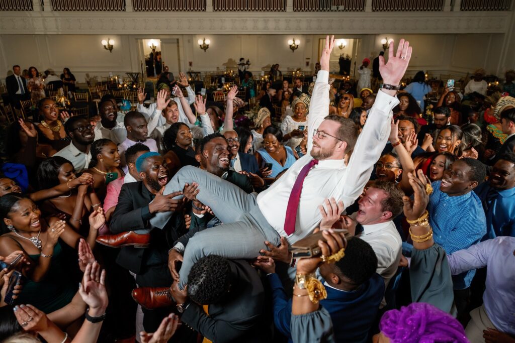 Groom with hands in the air while guests lift him in the air