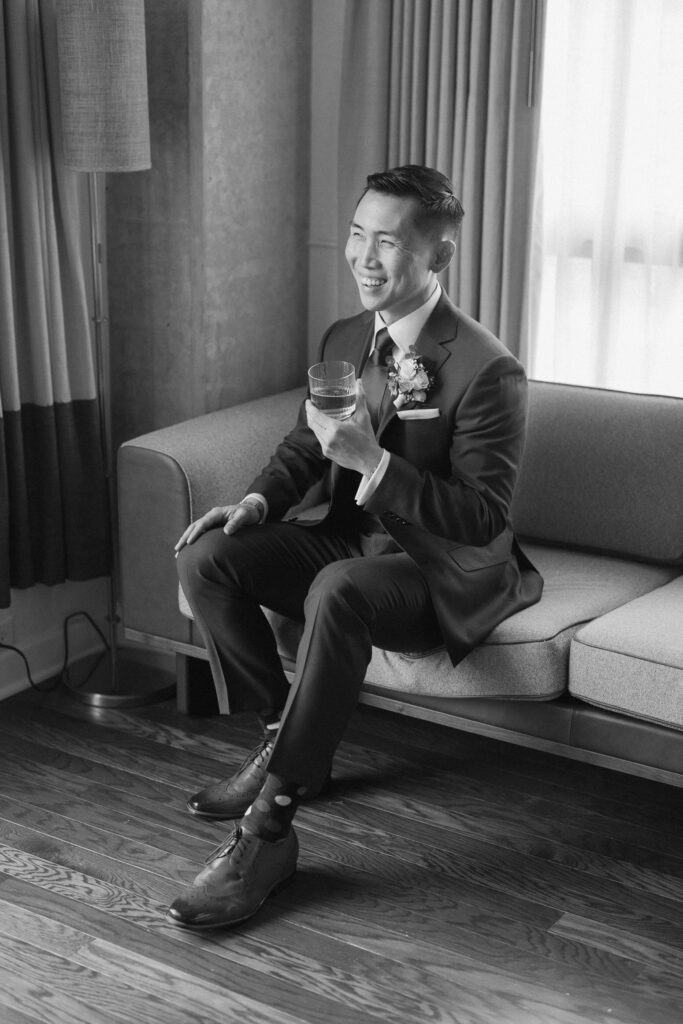 Groom sitting on couch in wedding attire holding glass cup with drink while laughing
