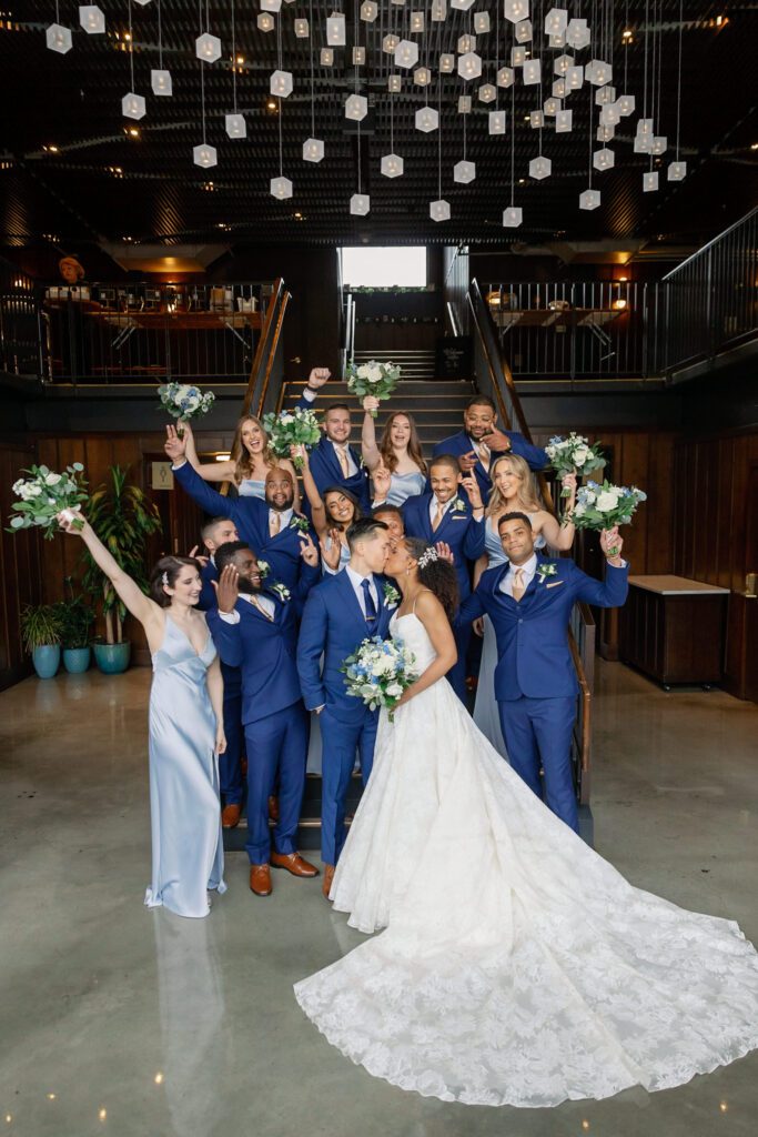 Couple kissing while bride holds bouquet and wedding party cheers behind them