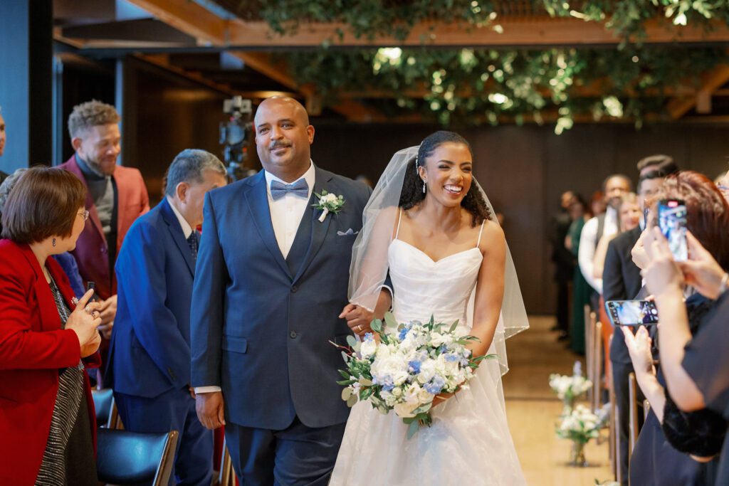 Bride smiling while walking down aisle with father holding bouquet